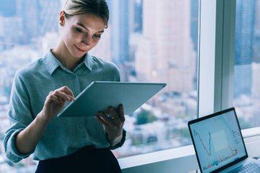 Modern ofiste çalışırken şehir planına karşı büyük pencerenin yanında bilgisayar masası olan çağdaş zarif bir kadın kablosuz dokunmatik ped kullanıyor.