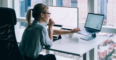 Gözlüklü şık sarışın kadının yan görünüşü. Çağdaş ofiste, büyük pencereli masada otururken dizüstü bilgisayar kullanıyor.