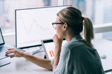 Modern ofisteki masaüstünde oturup istatistiksel grafikler izleyen gözlüklü çağdaş sarışın kadının yan görüntüsü