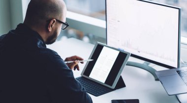 Büyük pencereli çağdaş ofiste dizüstü bilgisayar ve bilgisayarla oturup tablet izleyen gözlüklü kel işadamının yan görüntüsü