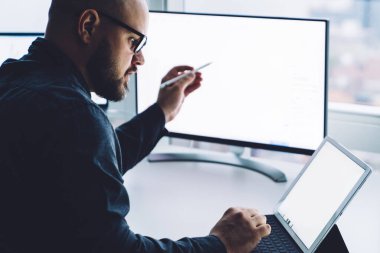 Modern ofiste pencereye karşı bilgisayarlı tablet kullanan gözlüklü yetişkin sakallı adamın yan görüntüsü