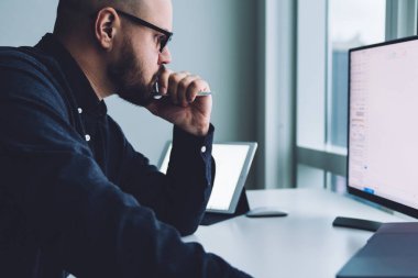 Ofisteki masasında oturan, bilgisayar kullanan ve grafik verileri üzerinde düşünen gözlüklü yetişkin bir adamın yan görüntüsü.