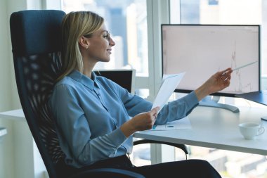 Modern ofisteki bilgisayar masasında çalışan güzel, kendine güvenen bir iş kadınının elinde belgelerle bilgisayarın monitörüne bakarken,