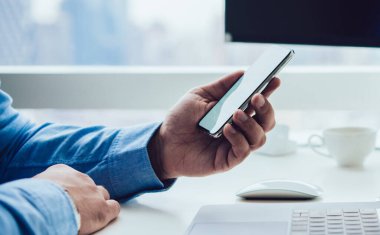 Ofisteki pencereye karşı dizüstü bilgisayarla masada otururken cep telefonu izleyen modern iş adamının yüzü olmayan yan görüntüsü
