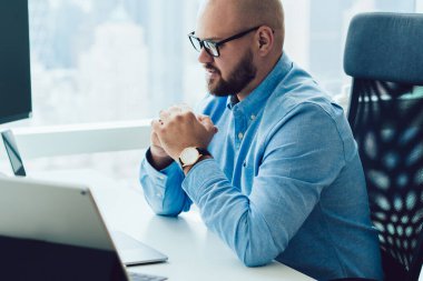 Gözlüklü ve mavi gömlekli kel adam el ele tutuşup modern ofiste bilgisayar masasında oturup dizüstü bilgisayar izliyor.