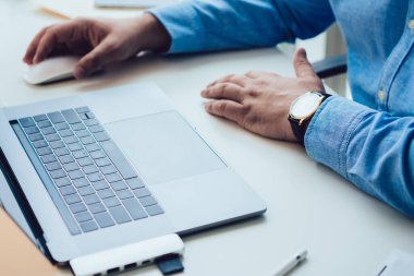 Mavi gömlekli yetişkin bir adam beyaz bilgisayar masasında oturuyor ve modern ışık ofisinde çalışırken dizüstü bilgisayar kullanıyor.