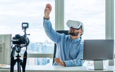Yetişkin bir adam ofiste bilgisayarlı ve şehir manzaralı bir masada oturuyor ve tripodla kameranın önünde sanal gerçeklik gözlüklerini kullanıyor.