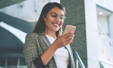 Zarif ceket giymiş neşeli bir Türk kadını, komik içerikli e-posta ile gülümsüyor, modern cep telefonlu mutlu bir kadın web sitesinde mesajlaşırken kendini iyi hissediyor.