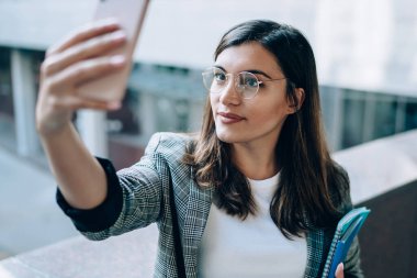 Modern cep telefonunun ön kamerasıyla selfie fotoğraflarına tıklayan şık gözlüklü çekici yeniyetme kız, eğitim için ders kitabı olan güzel bir kadın uygulamayla fotoğraf çekiyor.