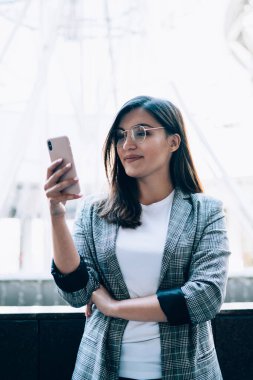 Gözlüklü olumlu kadın, modern cep telefonunda 4G kablosuz internete bağlı meslektaşlarıyla iletişim kurmak için yeni uygulama kuruyor, istatistikleri kontrol eden kadın blogcuları sorguluyor