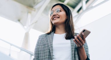 Sıradan ceketli mutlu genç bir kadının, şehir merkezinde telefon beklerken gülümsemesi, modern cep telefonunu elinde tutan, gözlüklü, pozitif hippi bir kızın görüntüsünün altında.
