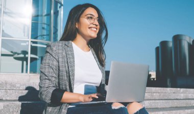 Modern bilgisayar cihazıyla şehir ortamında otururken mutlu bir metin yazarı gözlerini kaçırıyor ve gülümsüyor. Gözlüklü pozitif hippi kız dışarıda öğrenim görüp kendini iyi hissediyor.