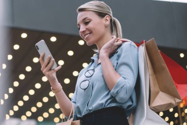 Telefon numarasını akıllı telefondan arayan neşeli genç bir kadın, açık havada 4G internet kullanıyor, cep telefonu tutan pozitif gülümseyen hippi kız ve kağıt torbalarla sokakta dururken uygulamayı güncelliyor.