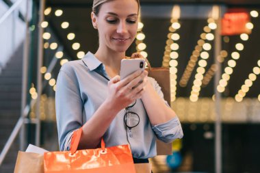 Alışveriş çantalı, modaya uygun hippi kız satış mağazalarında fiyatları kontrol etmek için akıllı telefon aygıtı kullanıyor. Reklam alanında 4G kablosuz bağlantıları var. Uygulama üzerinden mutlu beyaz kadın sohbeti.