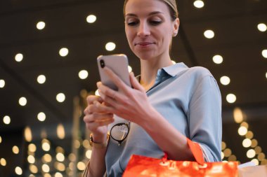 Modern akıllı telefonu ve kese kağıdıyla alışveriş saatinden sonra mesajları ve ihbarları karıştıran olumlu genç bayan alışverişçi, beyaz hippi kız alışveriş merkezleri hakkında bilgi arıyor.