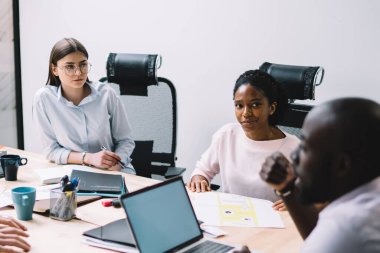 Yeni tasarım projesinde işbirliği yapan çok kültürlü erkek ve kadın meslektaşlar modern ofis içlerinde brifing toplantısı yapıyorlar, profesyonel iş adamlarından oluşan bir grup masada fikirleri tartışıyor.