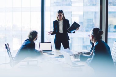 Şık takım elbiseli kafası karışmış kadın yönetmen toplantı odasında çalışanlarla iletişim kuruyor modern teknolojiyle toplantı masasının yanında duruyor, web sitesi için bilgisayarı var.