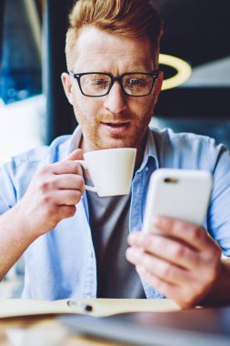 Genç adam rahat giyim tutarak fincan lezzetli kahve giymiş ve Web sitesinde 4g internet kullanarak akıllı telefon ile okuma haberleri, hipster Blogger zevk kafein içecek ve sosyal ağda video izlerken