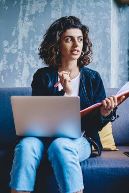 Şık saç kesimi olan düşünceli genç bir kadın elinde not defteri tutarken gözlerini kaçırıyor ve dizüstü bilgisayarla birlikte kanepede oturup yayıncılık hakkında yaratıcı fikirler düşünüyor.