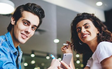 Gündelik giyinmiş neşeli erkek ve kadının portresi kafe 'de birlikte otururken kameraya gülümseyen ve selenlere mobil uygulama takan mutlu çift kahve dükkanında vakit geçiriyor.