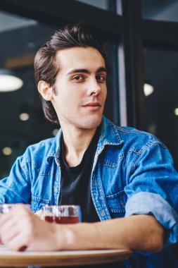 Kot ceket giymiş çekici bir gencin portresi kameraya bakarken modern kahve dükkanındaki akıllı telefon cihazını kullanan hippi kızla vakit geçiriyor.