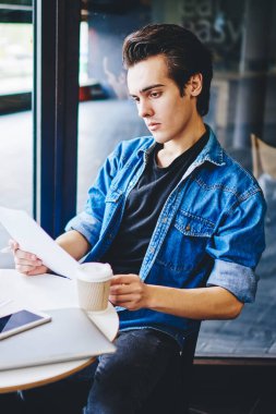 Düşünceli genç adam elinde kağıt belgeler tutarken ve bilgi okurken kız arkadaşı da akıllı telefon cihazıyla arayıp kahve dükkanının içindeki kahve masasında oturan ve not defterine notlar yazıyormuş.