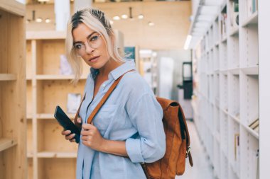 Kolejde kitaplıkların yanında elinde cep telefonuyla duran sıradan kıyafetli çekici bir kızın yarım boy portresi. Üniversite kütüphanesinde vaktini harcayan genç bir kız öğrenci.