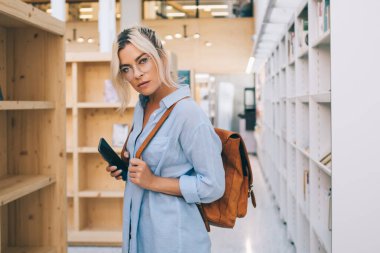 Kolejde kitaplıkların yanında elinde cep telefonuyla duran sıradan kıyafetli çekici bir kızın yarım boy portresi. Üniversite kütüphanesinde vaktini harcayan genç bir kız öğrenci.