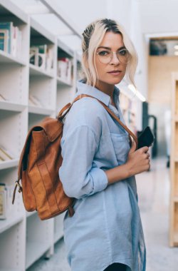Kolej kütüphanesinde rafların yanında duran optik gözlüklü beyaz kız portresi. Elinde akıllı telefon cihazı, sırt çantasıyla kameraya bakan genç bir kız öğrenci.