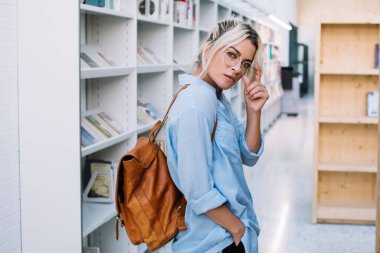 Okul kütüphanesinde eğitim için boş zamanlarını geçirirken kameraya bakan, kendine güvenen beyaz kadın öğrencinin yarım boy portresi. Sırt çantalı genç bir kadın.