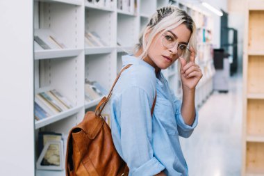 Okul kütüphanesinde eğitim için boş zamanlarını geçirirken kameraya bakan, kendine güvenen beyaz kadın öğrencinin yarım boy portresi. Sırt çantalı genç bir kadın.