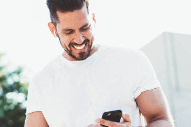 Neşeli adam gülerken içerik web sitesinde modern cep telefonunda gezinen internete bağlı komik video blogu izliyor, boş tişört giymiş mutlu bir erkek pozitif müzik listesinin keyfini çıkarıyor.