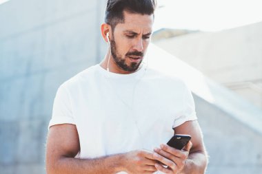 30 'lu yaşlardaki kafası karışmış bir adam, dışarıda dikilip, elektronik kulaklıkla yeni bir parça listesi dinlerken iş arkadaşından gelen e-postaları kontrol ediyor. Ciddi tişörtlü, sahte marka etiketli, SMS okuyan bir erkek.