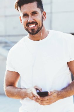 Boş tişört giymiş mutlu bir adamın yarım boy portresi kameraya bakıp gülümserken elektronik kulaklıklar ve mobil uygulamalarla yeni en çok satanlar listesine girmek için boş vakitlerini harcıyor.