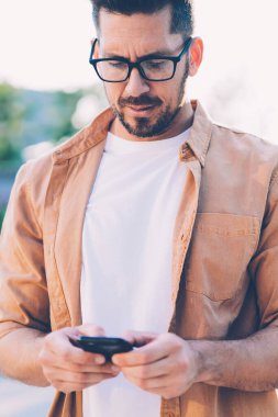 Gözlük takan ciddi bir erkek blog yazarı, akıllı telefon aygıtı ve açık havada 4G internet kullanarak e-posta mesajı yazıyor, sosyal ağda cep telefonuyla haber okuyan tişörtlü bir adam