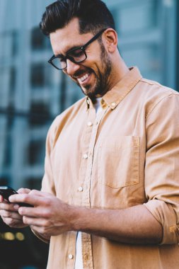 Gözlüklü başarılı sakallı adam, gözlüklü, göz koruyuculu, gülümseyen, arkadaşından pozitif mesajlar okuyan, 4G 'ye bağlı, mutlu erkek web sitesi akıllı telefon aygıtı ile dışarıda dururken,