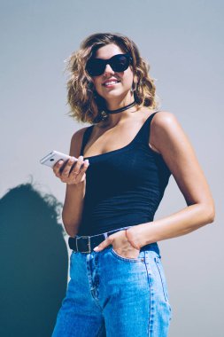 Akıllı telefon zımbırtısıyla kameraya bakan mutlu milenyum hipster kızının yarım boy portresi. Güneş gözlüklü pozitif bir kadın boş vakitlerinde elinde cep telefonu tutuyor.