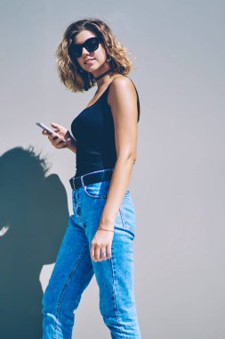 Şehir sokaklarında yürüyüş sırasında elle dijital gadget ile şık milenyal hipster kız yarı uzunlukta portre, kamera bakıyor güneş gözlüğü çekici kadın