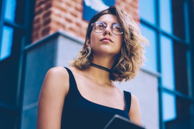 Trendy güneş gözlüklü düşünceli hipster kız arka planda üniversite ziyaret ettikten sonra kentsel ortamda dinlenirken dikkatli görünümlü, beyaz kadın öğrenci kolej zamanında durulması