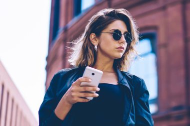 Akıllı telefon Gadget tutarak ve taksi hizmeti, trendy kadın turist harcama ücretsiz zaman seyahat ve telefon kullanarak çağrı beklerken uzak görünümlü güneş gözlüğü emin kadın