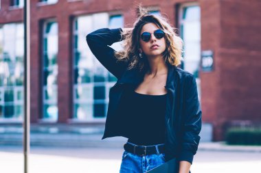 Üniversite kampüsünde elinde dosyayla gezen kendinden emin bir kız öğrencinin yarım boy portresi. Kameraya bakarken, beyaz, kot pantolonlu ve şık güneş gözlüklü çekici kadın.