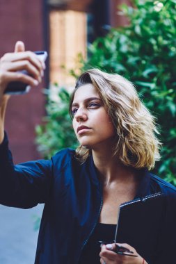 Üniversiteden dönerken akıllı telefon kamerasına bakarak selfie çeken genç ve güzel hippi kız. Mobil cihazla medya uygulaması üzerinden fotoğraf çekmek için poz veren ciddi bir kız öğrenci.