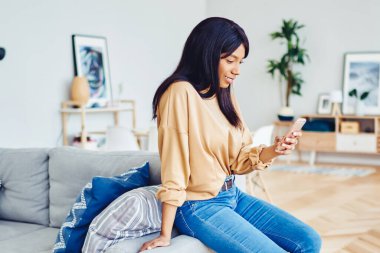 Genç Afrikalı Amerikalı kadın oturma odasında koltukta oturuyor boş zamanlarında 4G 'ye bağlı cep telefonu kullanıyor, koyu tenli hippi kız sosyal ağlardaki içeriği hücresel ağlarda paylaşıyor.