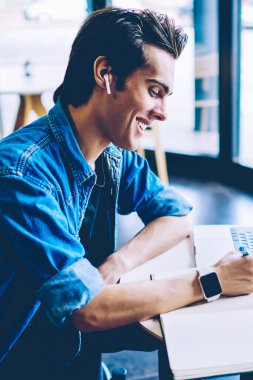 Heyecanlı gülümseyen hipster adam bluetooth elektronik kulaklıklar ile olumlu müzik çalma listesi dinlerken ders kitabında kompozisyon yazarken ve modern dizüstü bilgisayar ile öğrenme. Netbook bilgisayarda freelancer çalışma