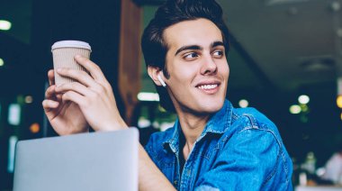 Neşeli hipster adam kulaklık ile olumlu ses kaydı zevk sırasında paket kahve tutan, genç gülümseyen adam müzik dinlerken ve dizüstü bilgisayarda iletişim mola uzağa bakıyor