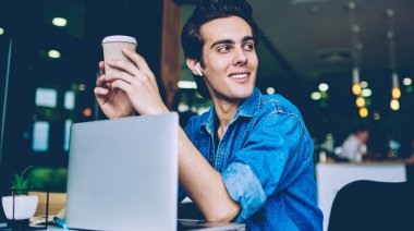 Neşeli hipster adam kulaklık ile olumlu ses kaydı zevk sırasında paket kahve tutan, genç gülümseyen adam müzik dinlerken ve dizüstü bilgisayarda iletişim mola uzağa bakıyor