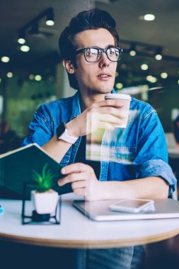 Sınava hazırlanmak için bilgi literatürü kullanarak kapalı mekanda kafeinli içecek keyfi gözlük genç adam Pondering, düşünceli hipster adam rahat paket kahve tutarak giyinmiş