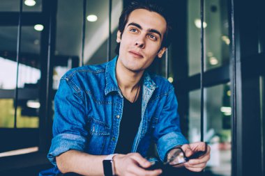 Rahat kıyafet giymiş hipster adam elinde optik gözlük tutan ve düşünceli serbest zaman sırasında uzağa bakarak, dalgın erkek öğrenci arkadaş beklerken üniversite sınavları hakkında düşünme