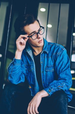 Ciddi erkek öğrenci kot giyim giyinmiş merdivenlerde oturan ve marka tasarımcıları hakkında düşünme hissi, vizyon düzeltme düşünceli uzak görünümlü gözlük düşünceli hipster adam