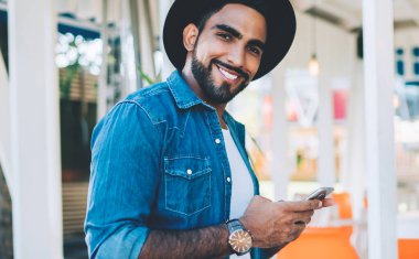 Yarım boy, şık siyah şapkalı İspanyol hippi portresi elinde cep telefonuyla halka açık bir yerde poz veriyor modaya uygun giyinmiş kameraya gülümseyen ve blog yazan bir adam.